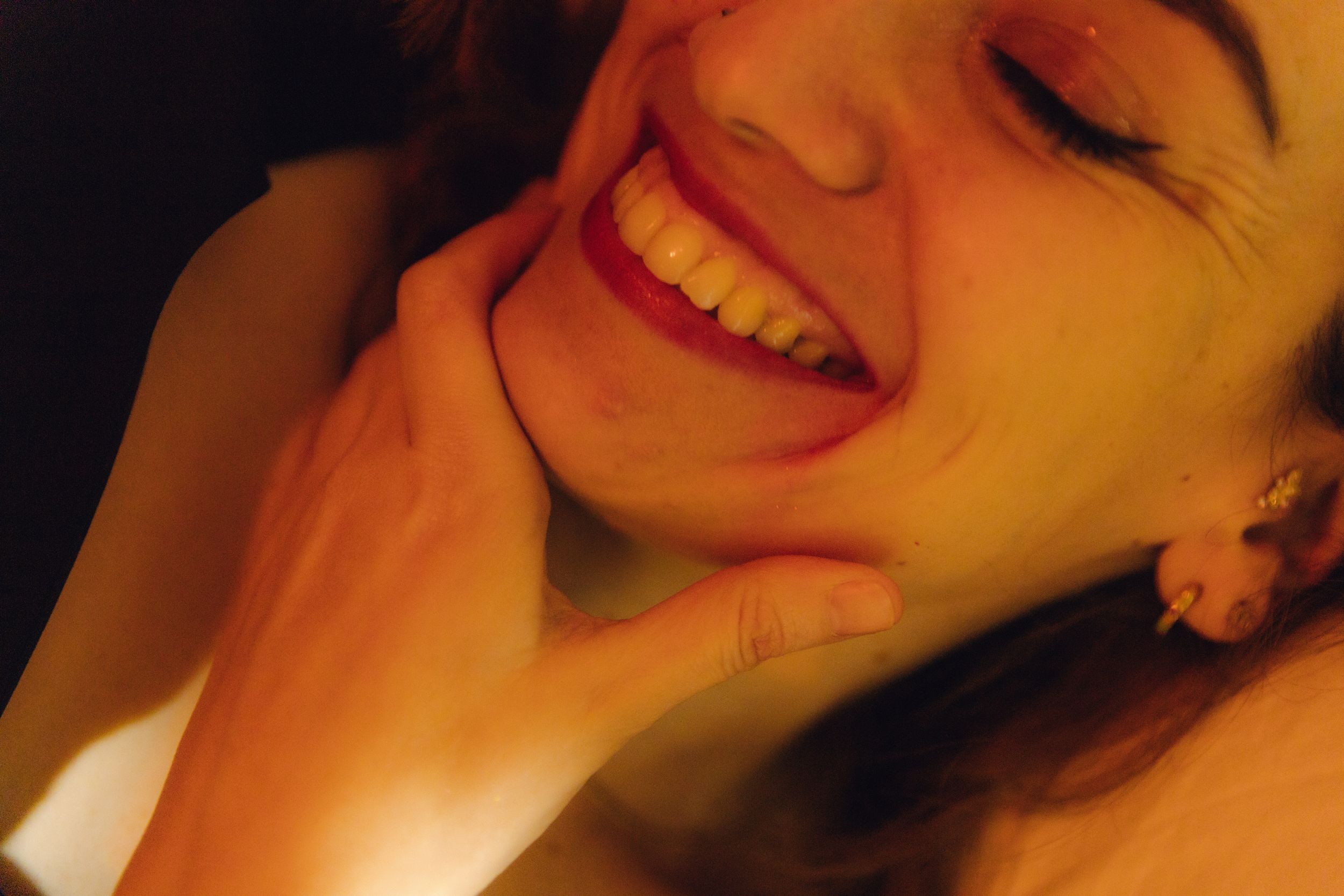 A young woman with red lipstick smiles joyfully while cradling her face in her hands in warm golden light.