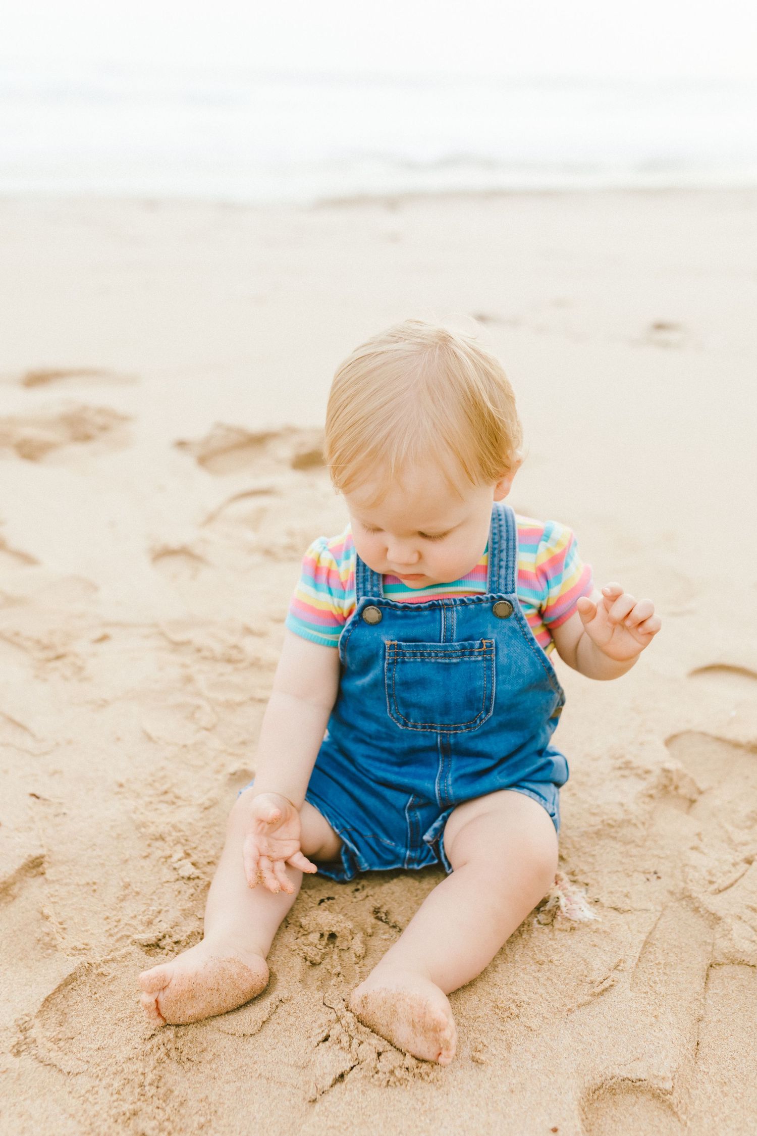 Small child in denim overalls plays in the sand at the beach during a sunny day.