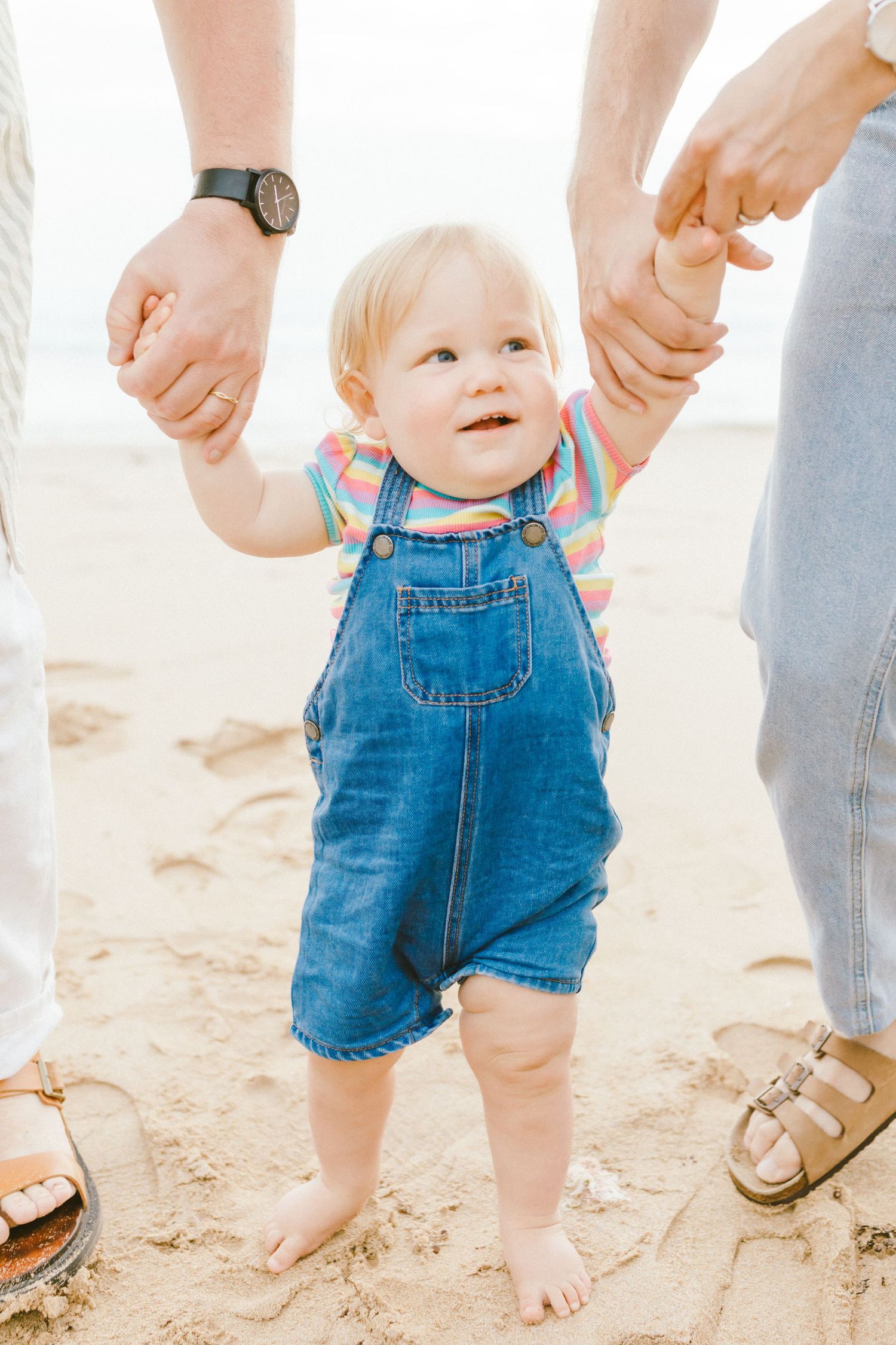 Small child in denim overalls takes first steps on sandy beach while holding parents' hands for support.