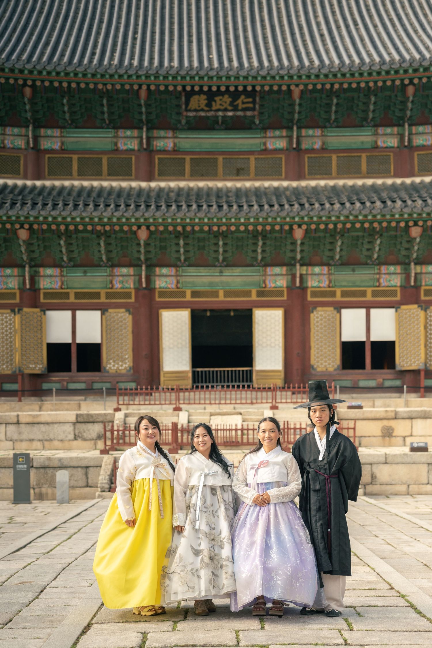 Group wearing traditional Korean hanbok dresses poses in front of historic Gyeongbokgung Palace architecture.