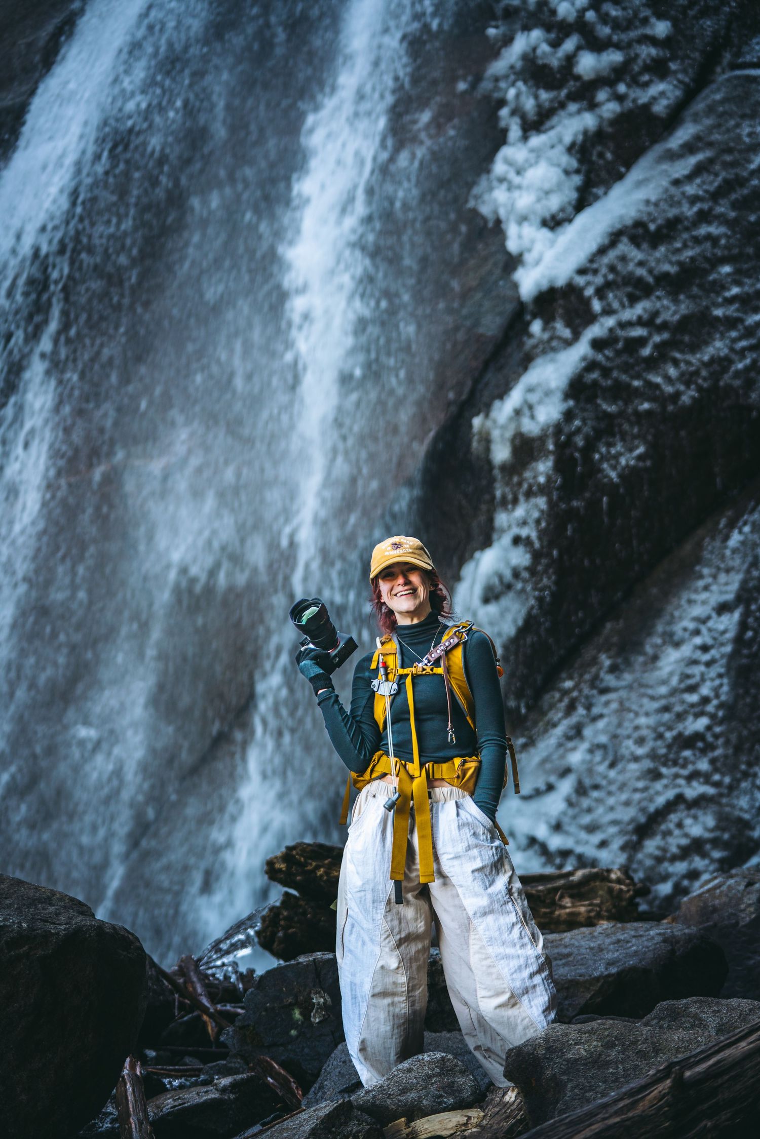 Female photographer in white pants and black shirt holding a camera in front of a stunning waterfall.