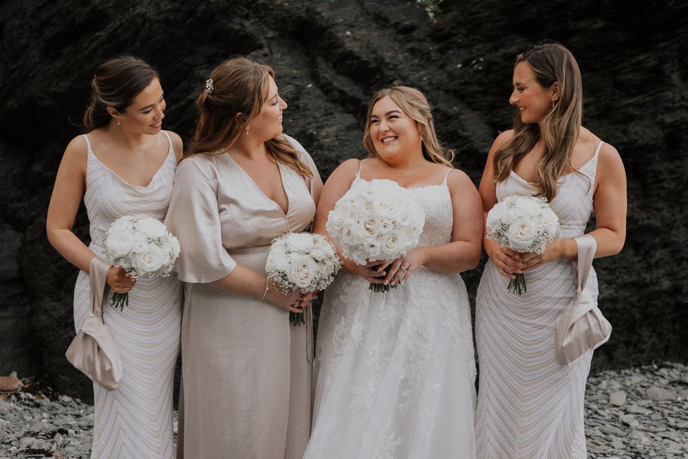 Bride with three bridesmaids in champagne dresses holding white bouquets on rocky beach.
