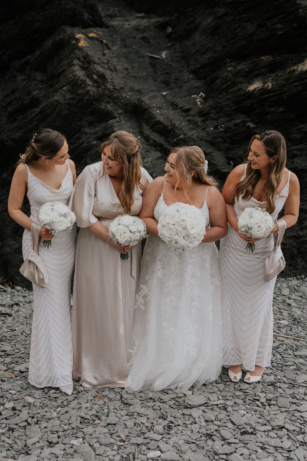 Bride and three bridesmaids in champagne gowns holding white bouquets on pebble beach.
