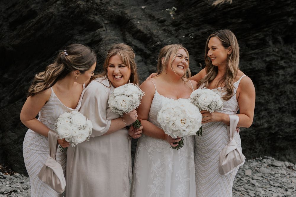 Bride and three laughing bridesmaids in champagne dresses holding white bouquets on rocky beach.