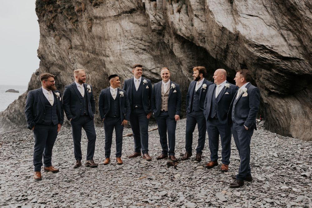 Groomsmen in navy suits posing on a rocky beach with dramatic cliff backdrop.