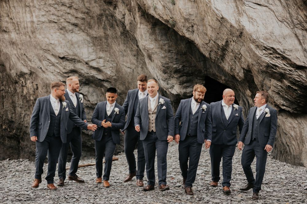 Wedding party groomsmen in navy suits walking and laughing on a pebble beach.