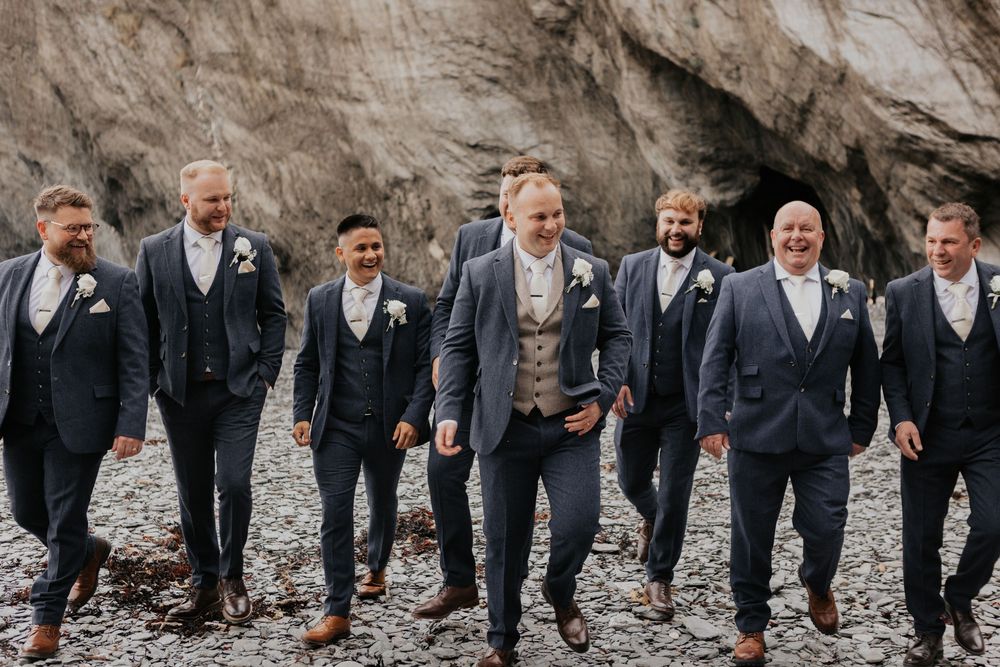 Groom and groomsmen in navy suits walking confidently on a rocky coastal beach.