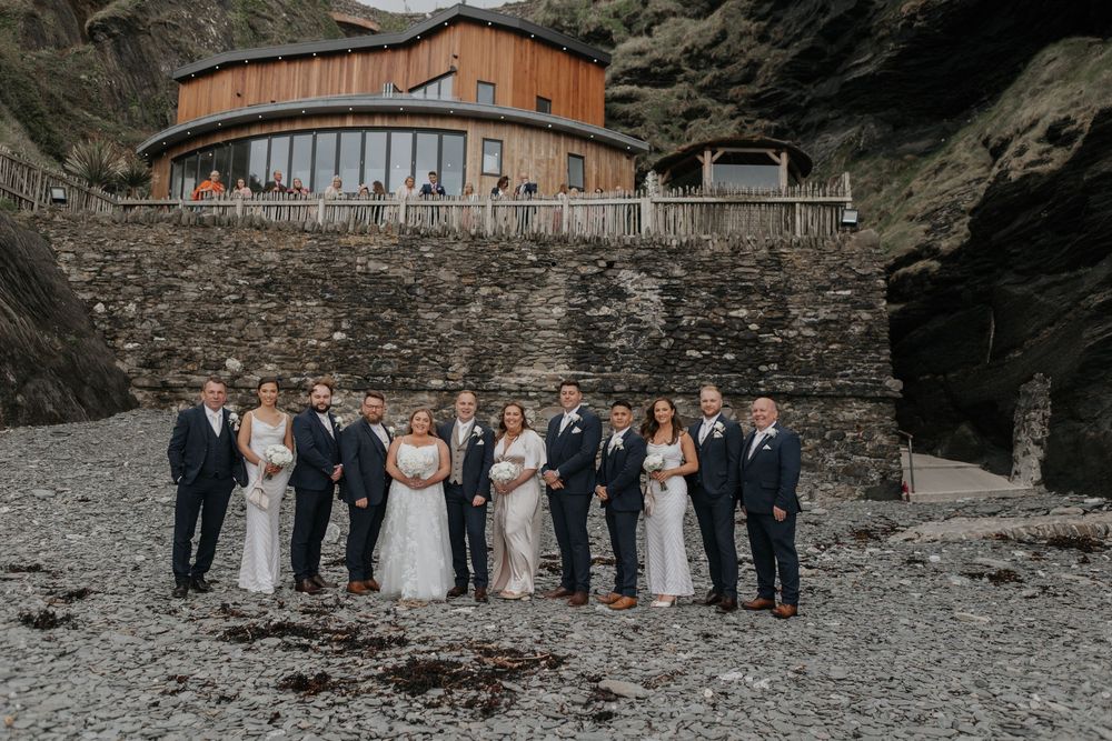 Full wedding party posing on a rocky beach in front of a circular wooden venue.