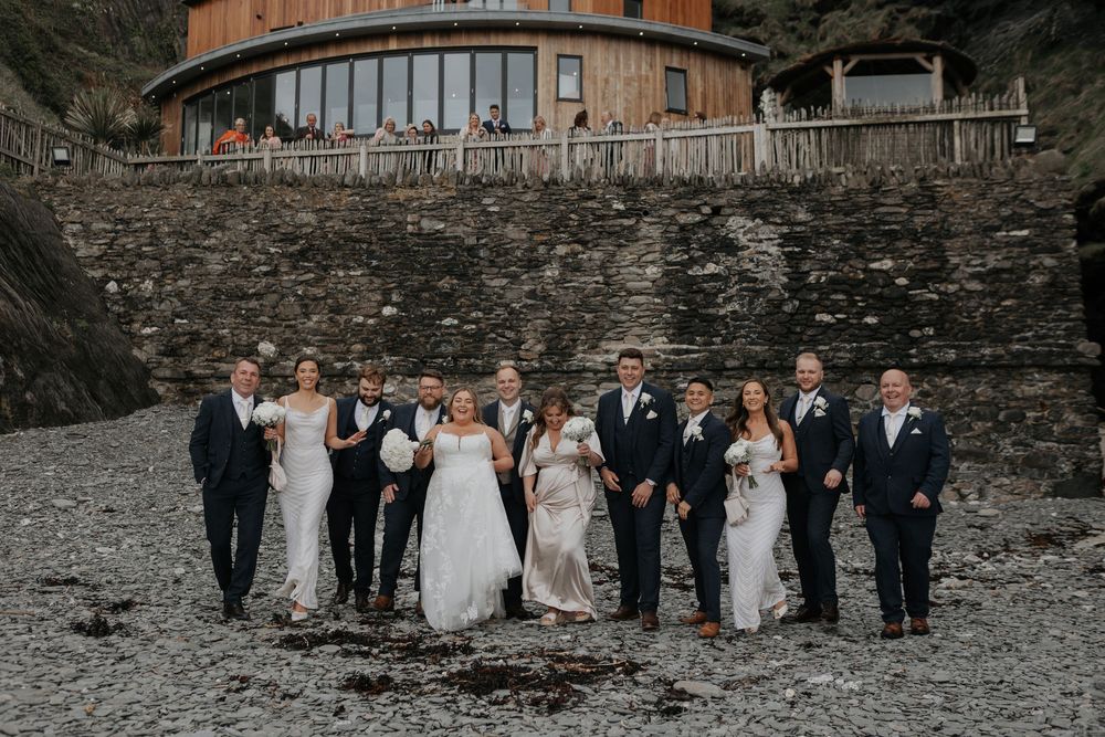 Bride, groom, and wedding party walking on a pebble beach with coastal venue behind.