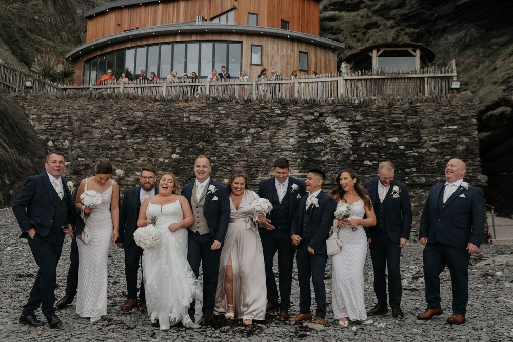 Wedding party laughing and posing on a rocky beach with a wooden circular building.