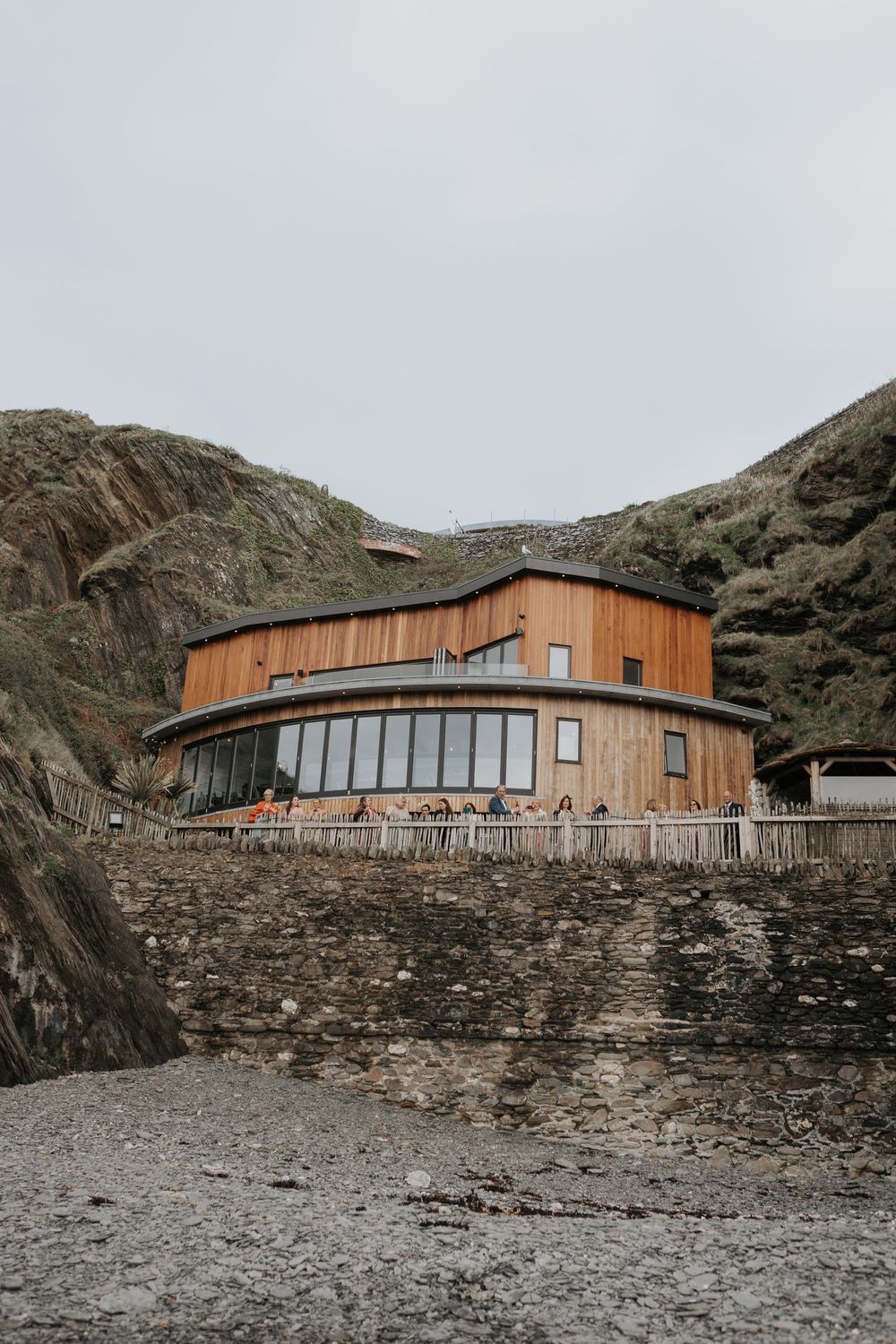 Circular wooden coastal wedding venue nestled between rocky cliffs on an overcast day.