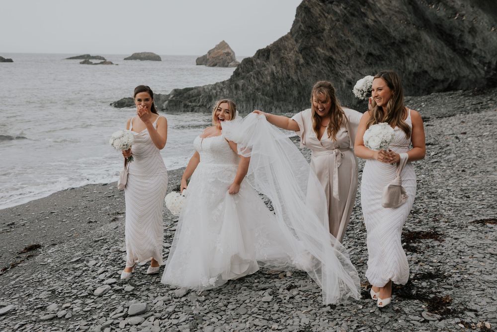 Bride and three bridesmaids in white dresses walking on a rocky beach by the sea.