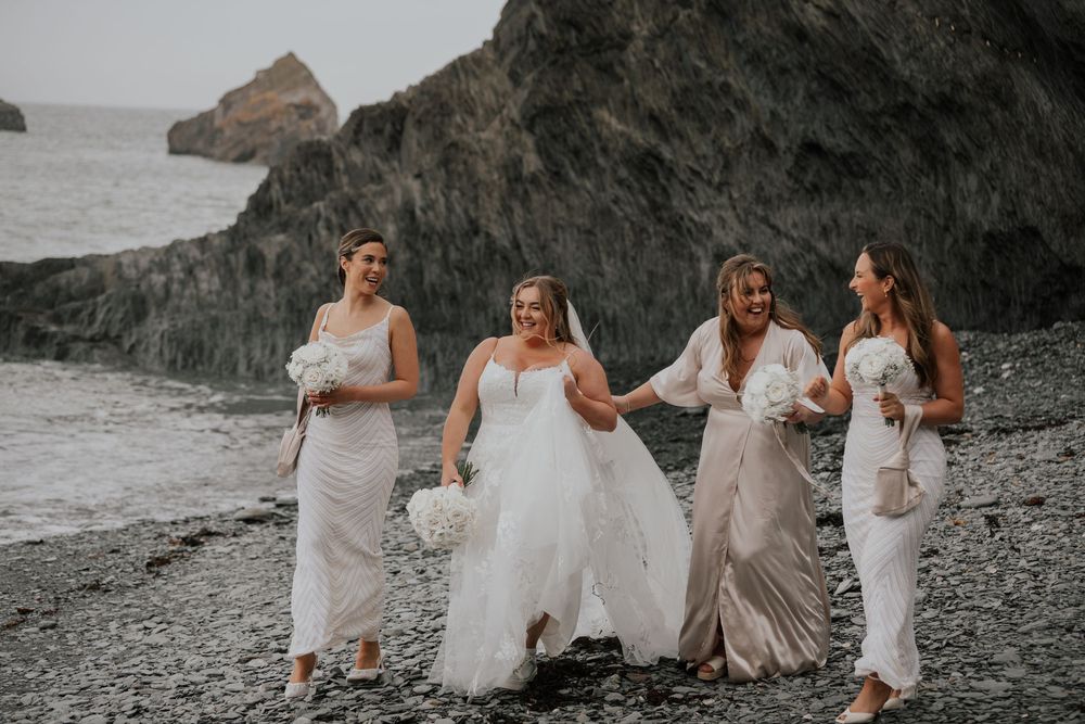 Bride in white gown and bridesmaids in neutral dresses strolling on a pebble beach.