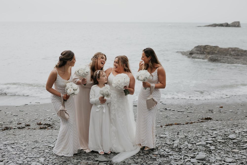 Bride and bridesmaids laughing together on a rocky beach holding white bouquets.