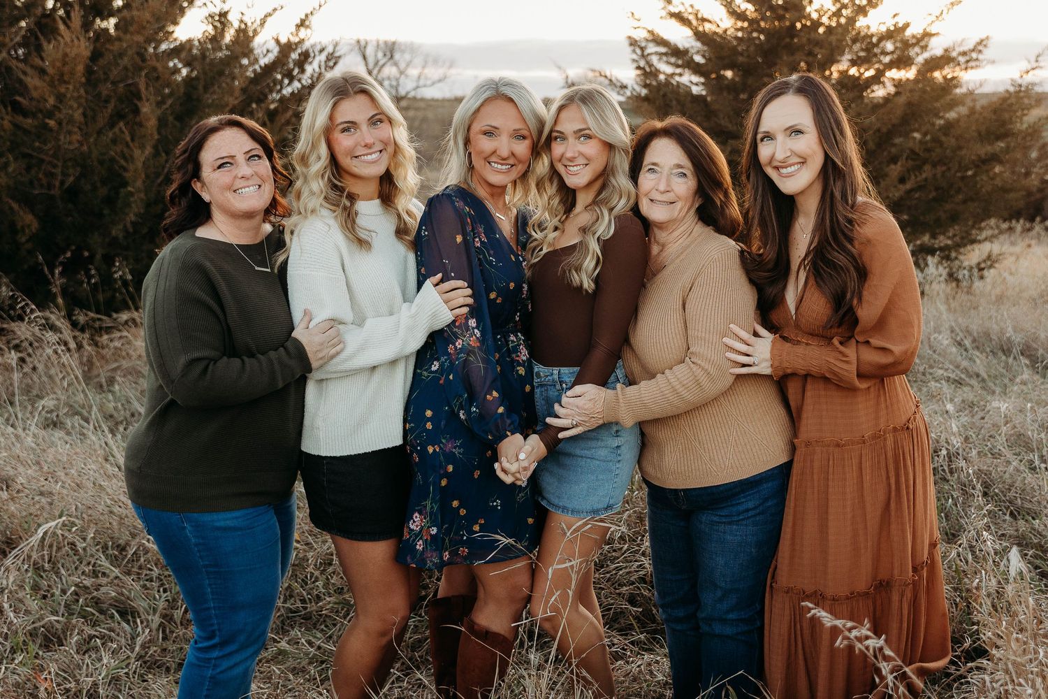 A group of people wearing casual fall attire pose together outdoors at sunset in a grassy field.