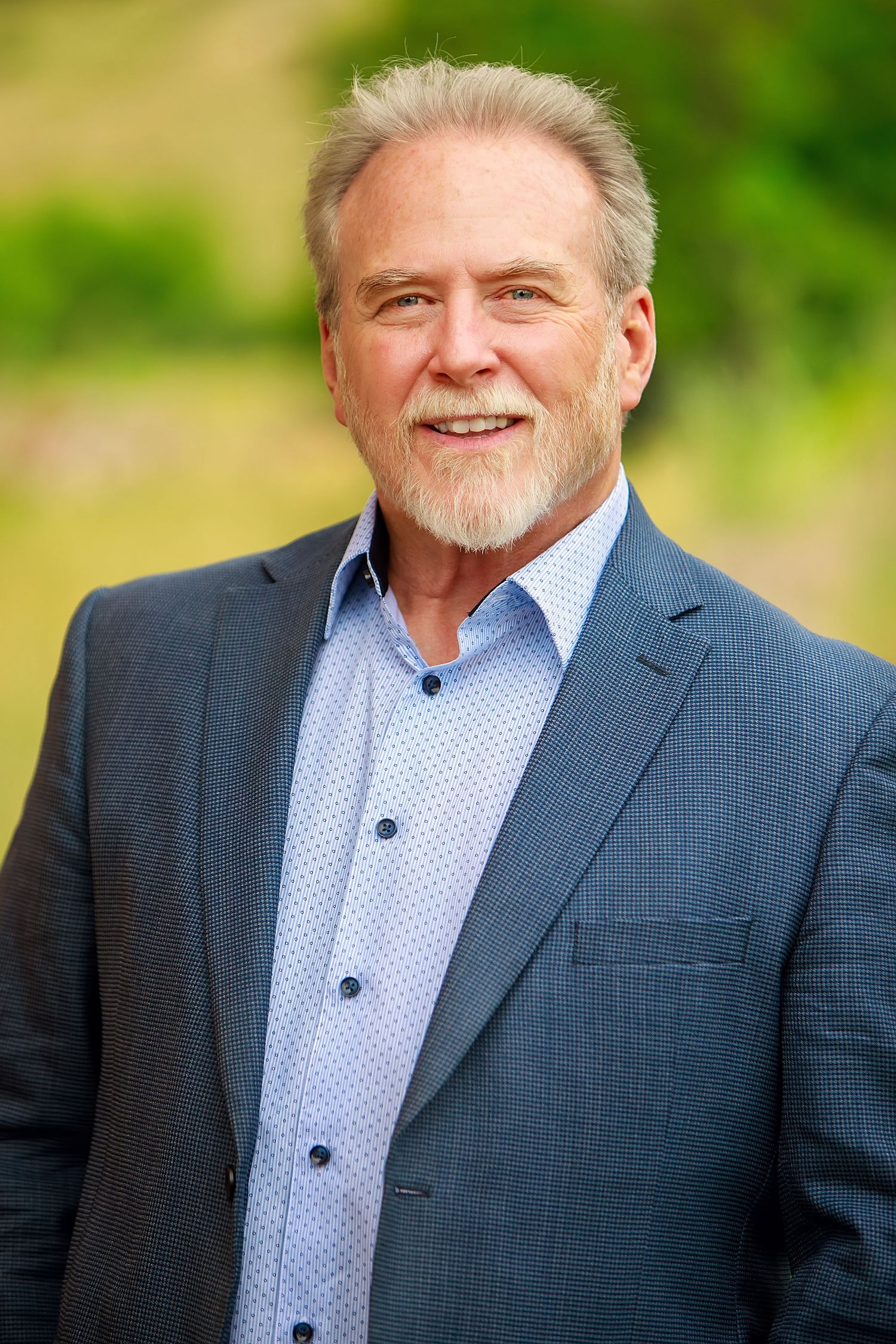 Professional headshot of mature man in blue suit with gray beard smiling outdoors.