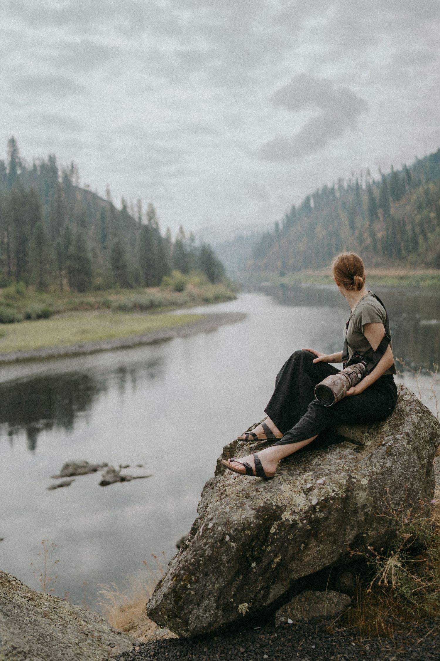 A person relaxes on a large boulder beside a misty river surrounded by evergreen forests in the Pacific Northwest.