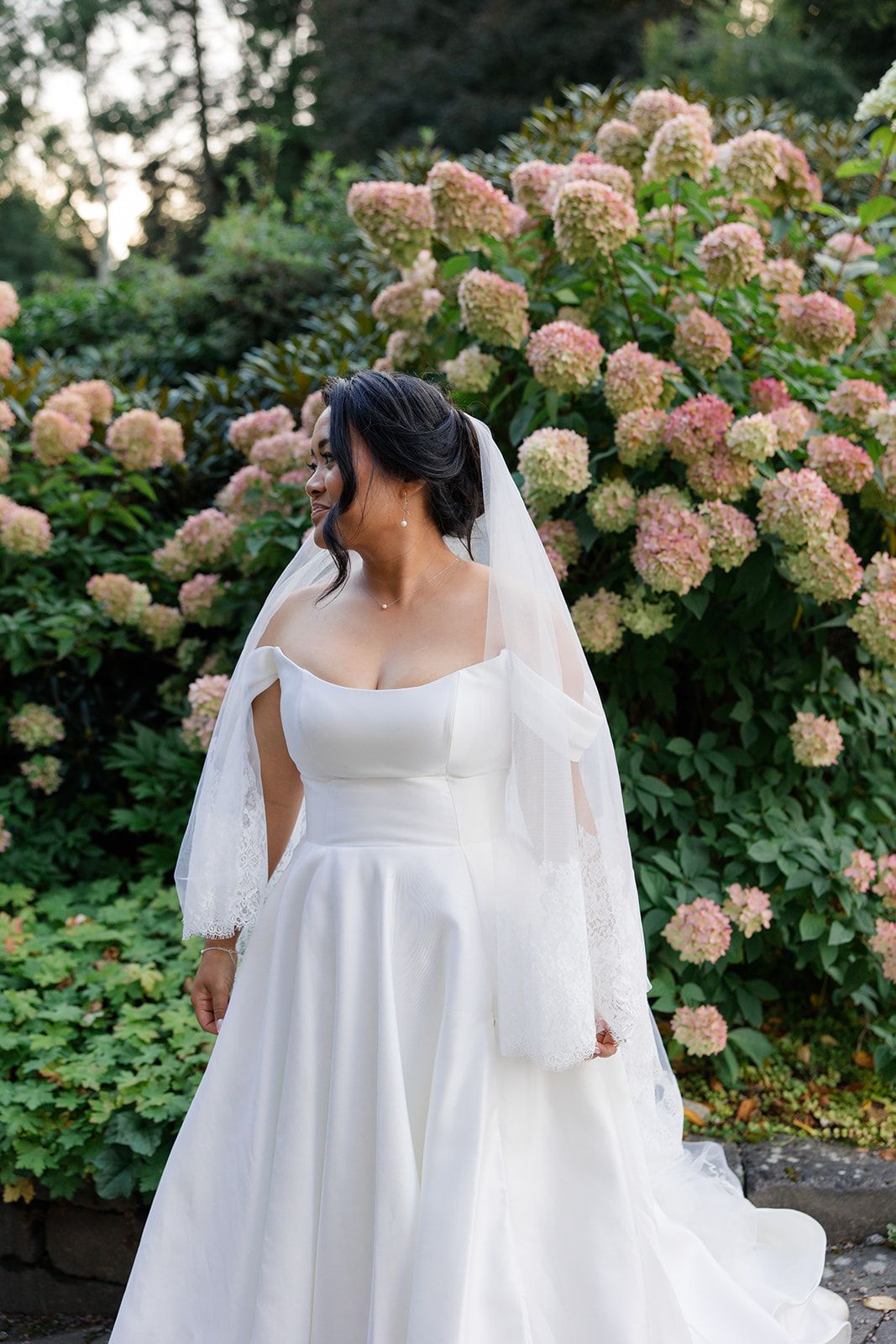 Two people in flowing white wedding attire stand together among blooming pink hydrangea bushes in a garden setting.