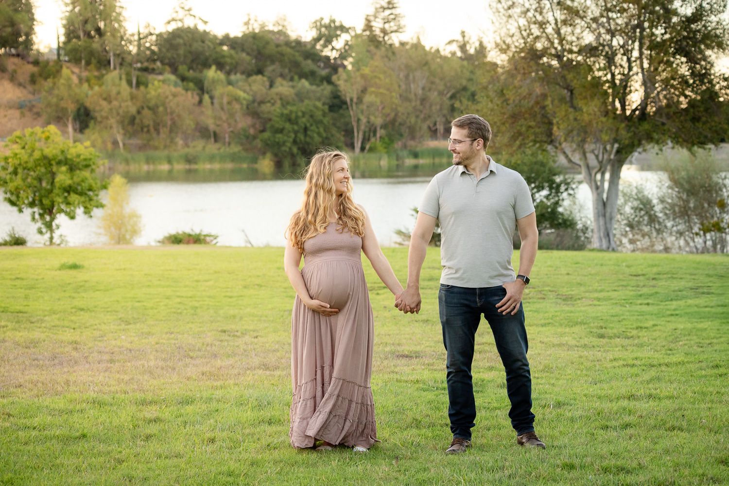 A couple expecting a baby holds hands while standing on grass near a tranquil lake during golden hour in the Bay Area