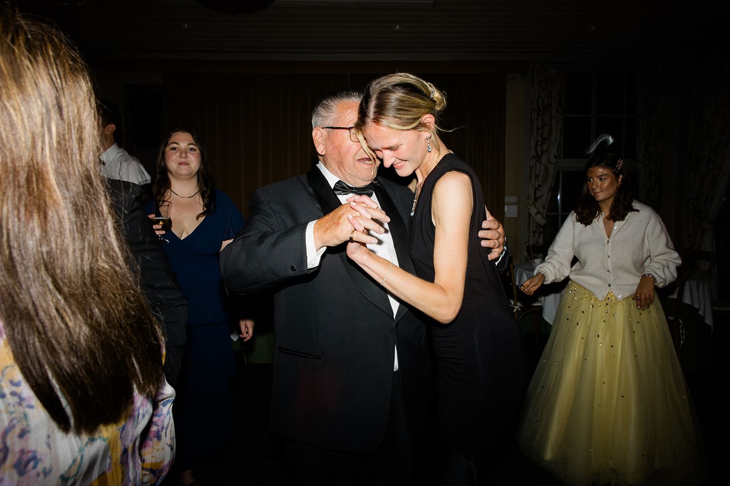 Couples dance together at a dimly lit reception venue while guests watch from the background.