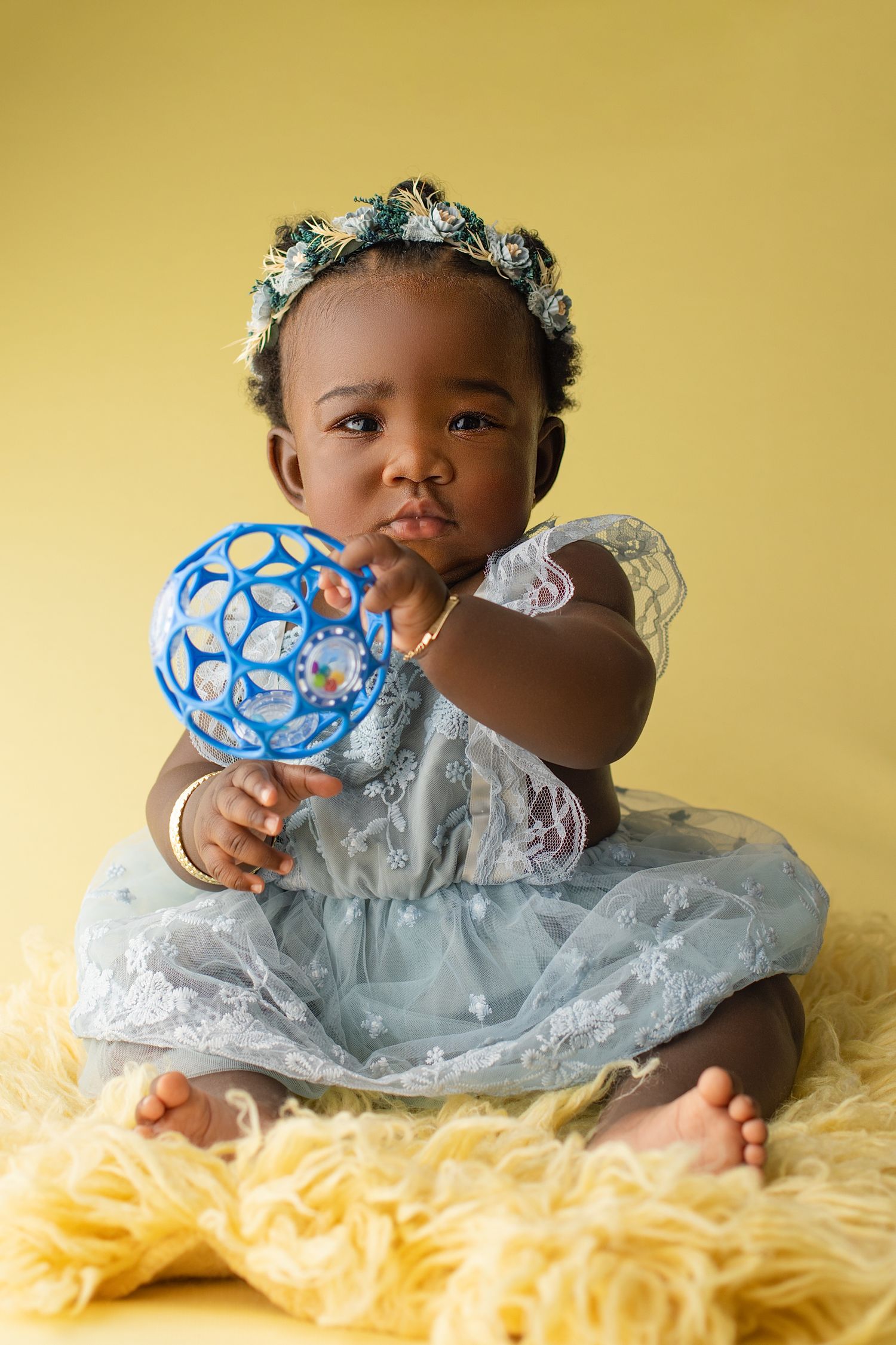 A small child in a light blue dress and floral crown holds a blue sphere toy while sitting on yellow fabric.