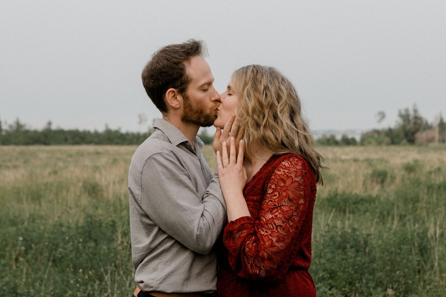 A couple in a field shares a romantic moment, with her wearing a red dress and him in a gray suit.