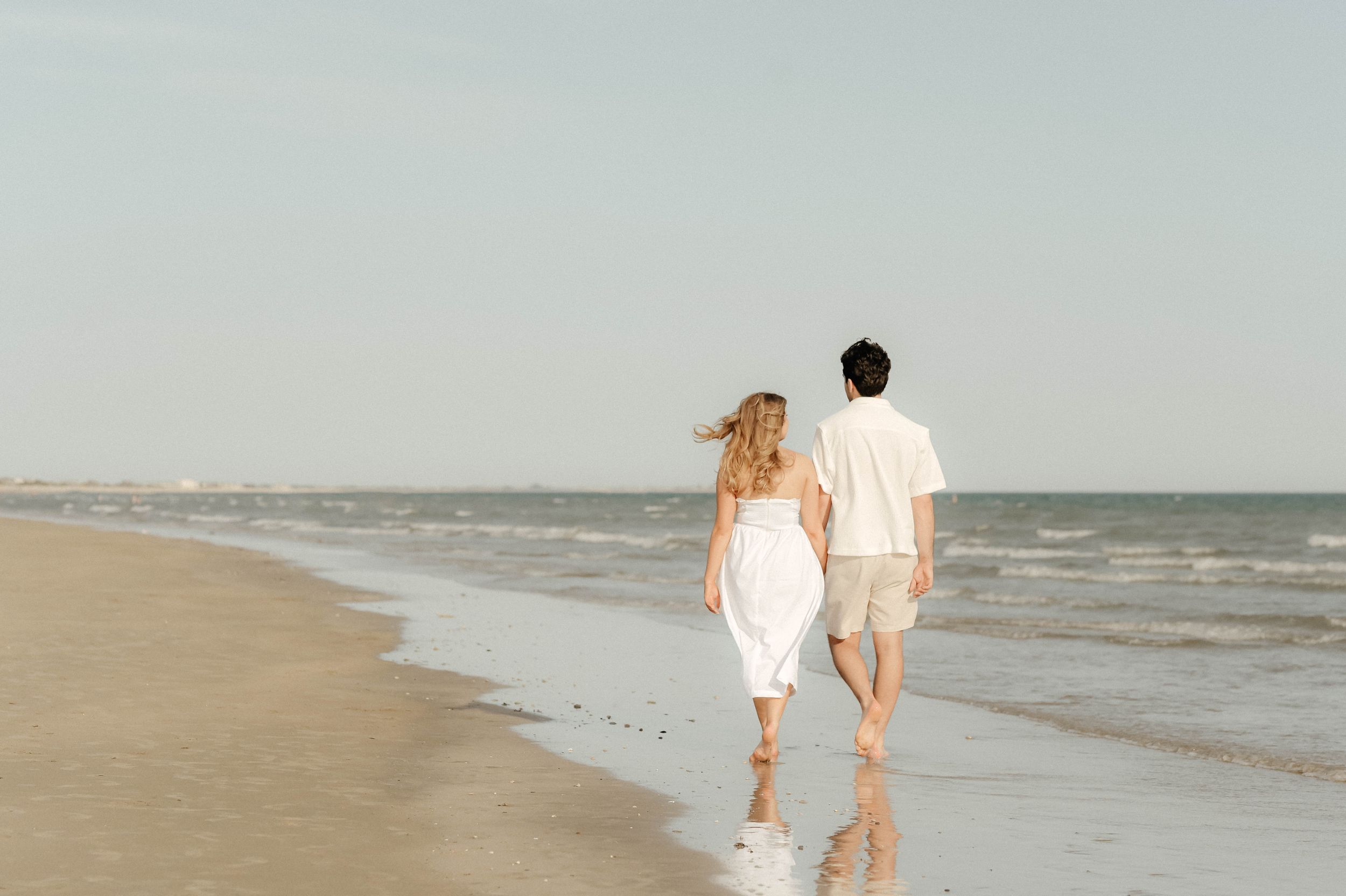 A couple in white outfits walks hand in hand along a serene beach shoreline during sunset.