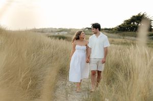 A couple in white outfits walks through a golden wheat field at sunset holding hands.