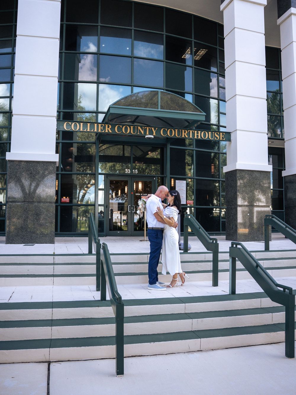 Three people stand on the steps of the Collier County Courthouse entrance on a sunny day in Naples, Florida