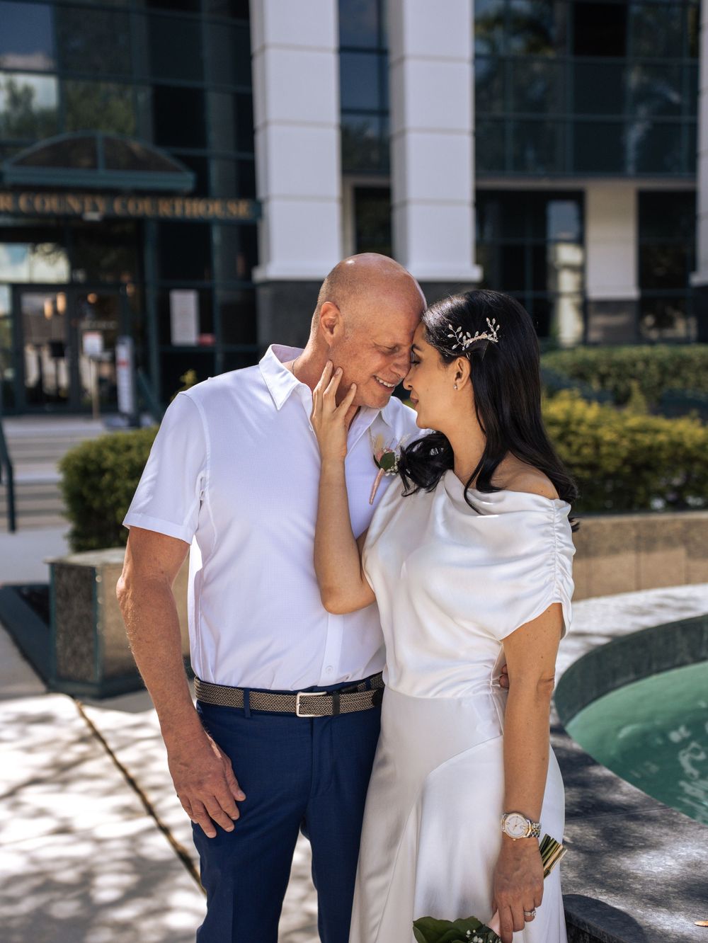 A couple shares a tender kiss while standing outdoors near a building with white columns and glass windows in Naples, Florida