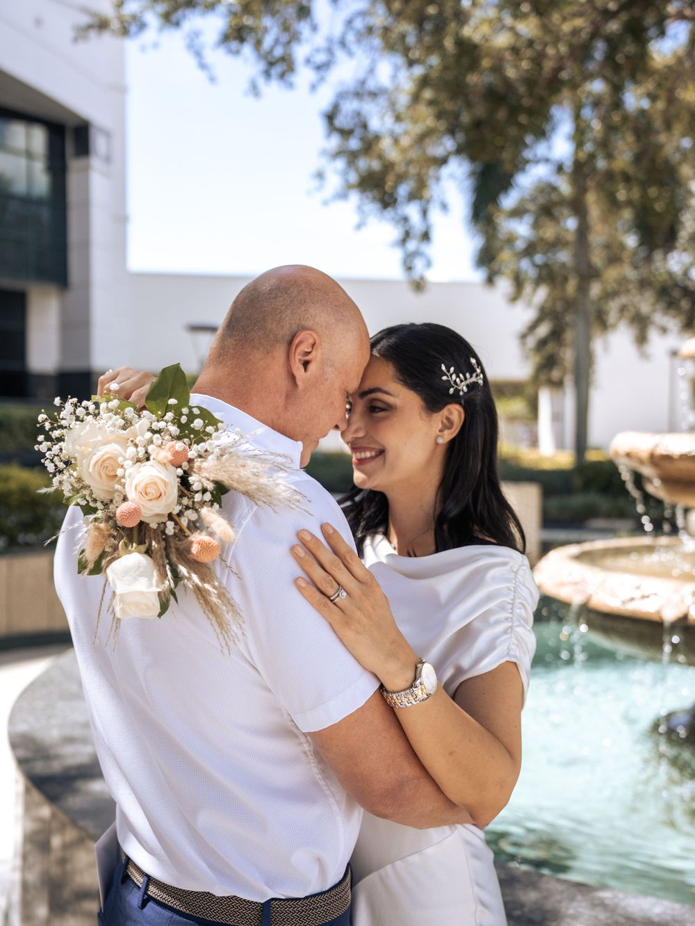 A couple in white shirts share tender moments by a pool on a sunny day with decorative floral arrangements in Naples, Florida