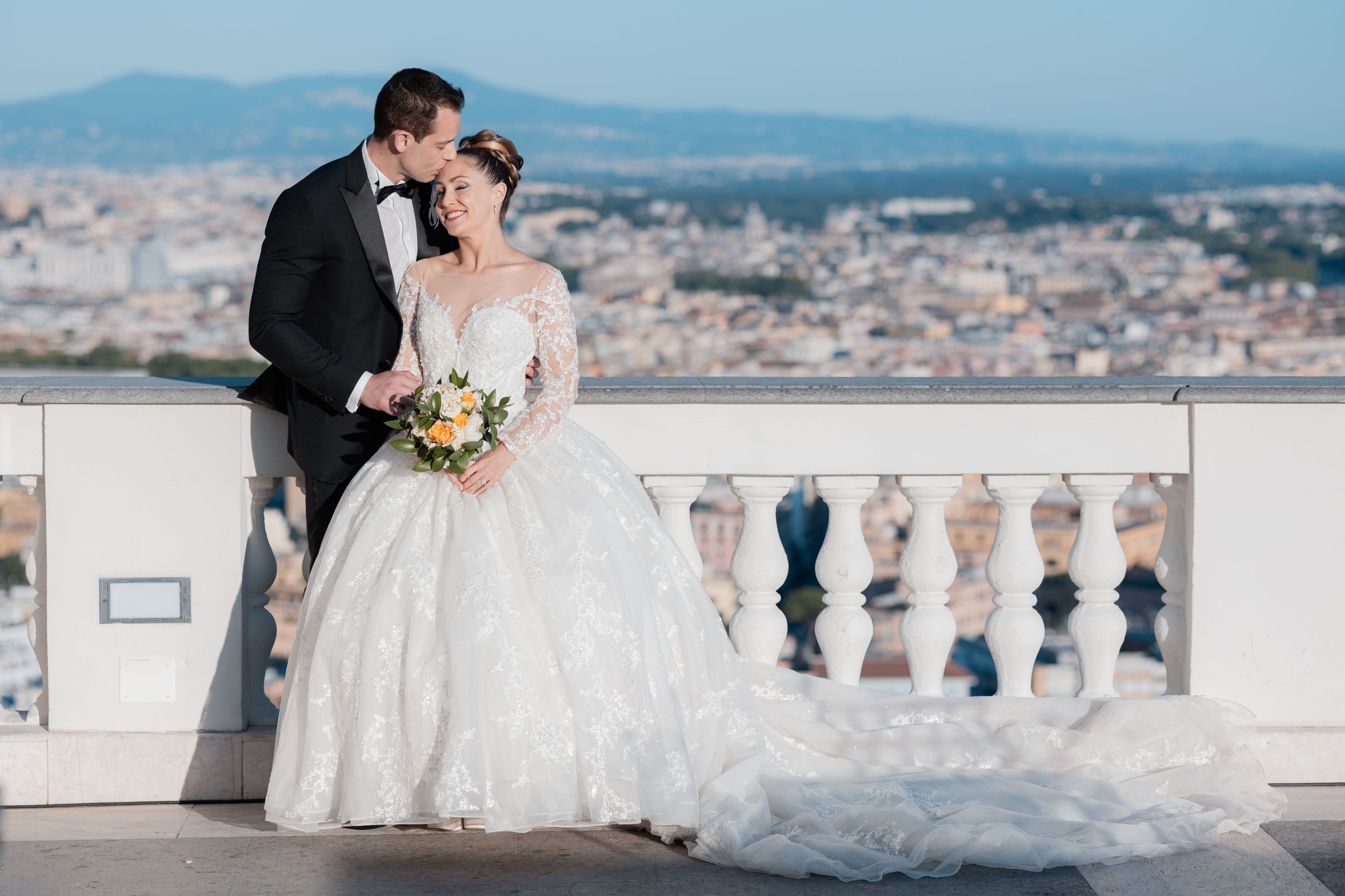 A romantic wedding couple stands on a balcony overlooking a scenic cityscape with mountains in the distance.