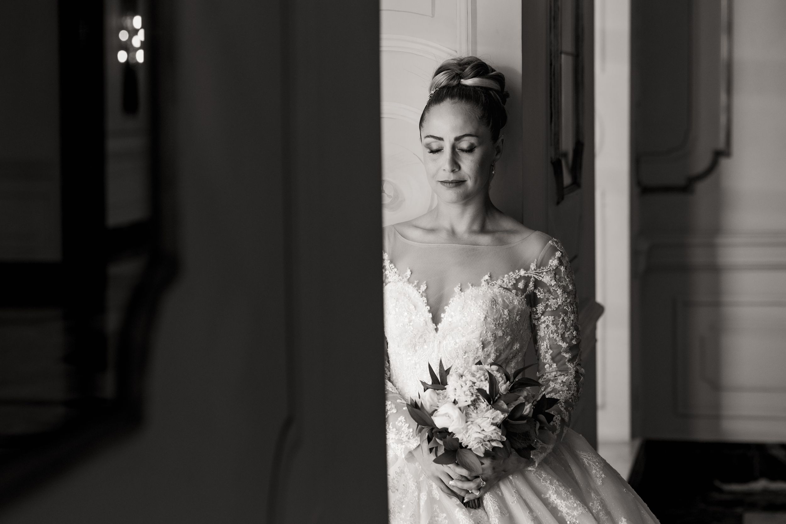 A bride in a white lace wedding gown stands in a doorway with dramatic lighting and shadows.