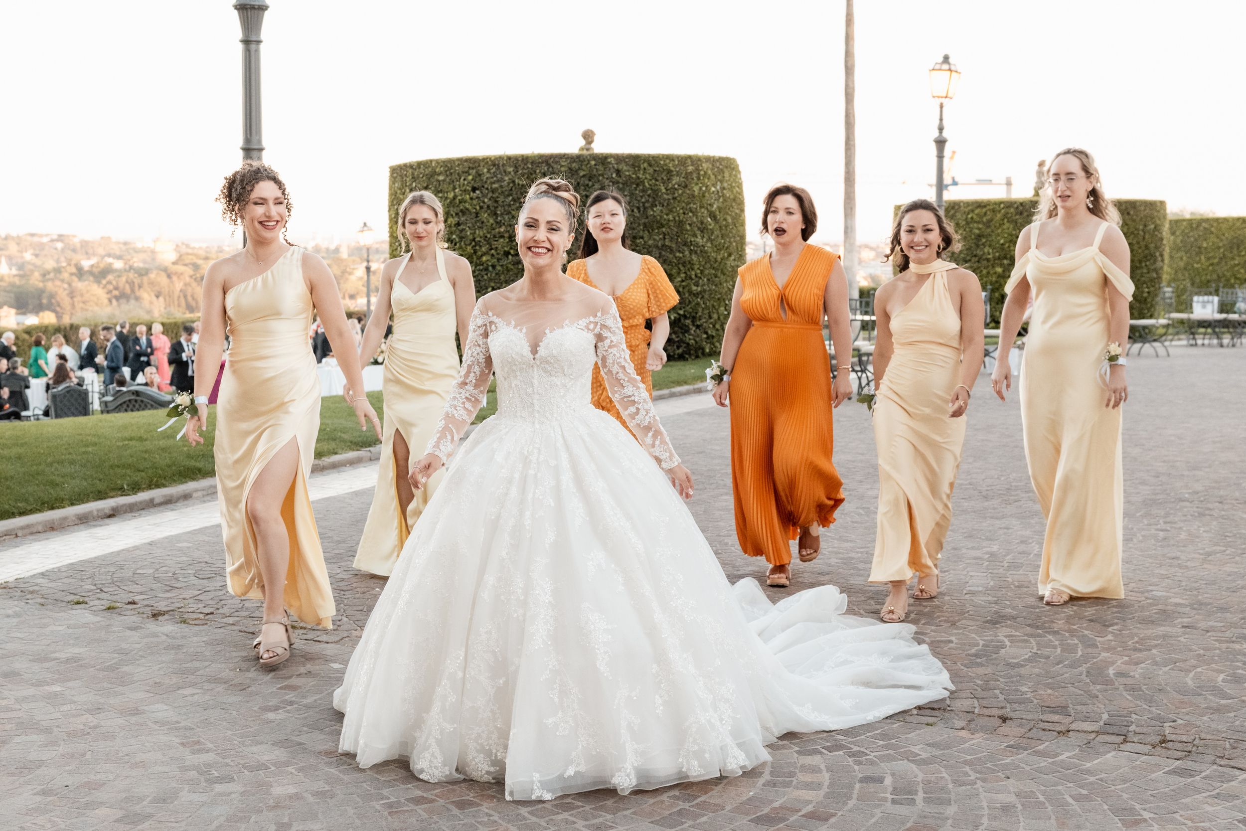 Wedding party wearing peach and orange dresses poses together on stone path during outdoor celebration.