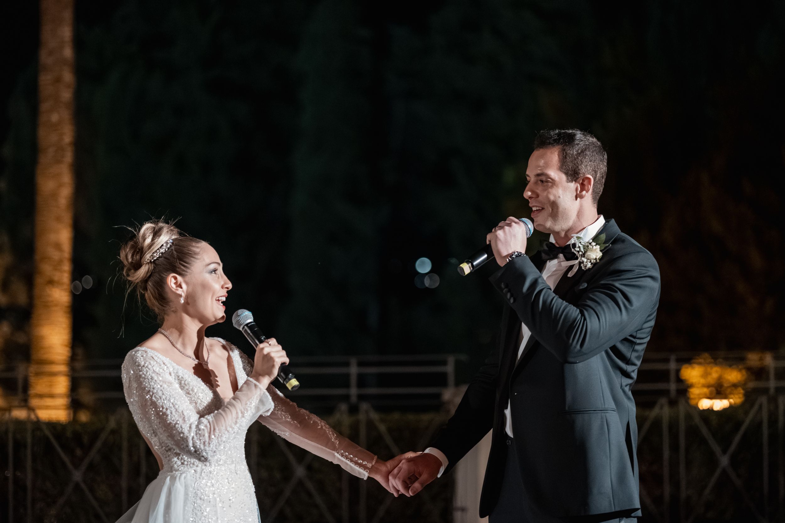 A couple performs a duet with microphones during their outdoor nighttime wedding celebration.