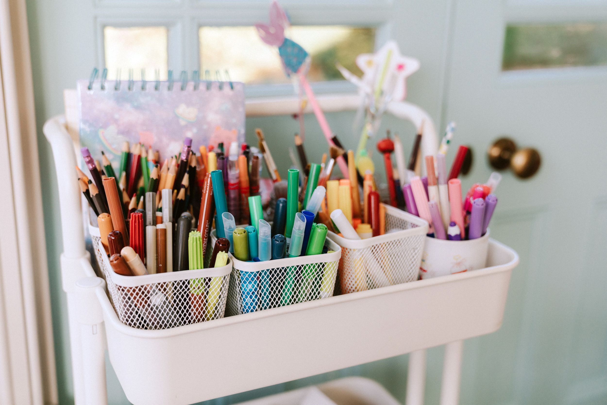 Colorful art supplies and markers organized in white containers on a shelf.
