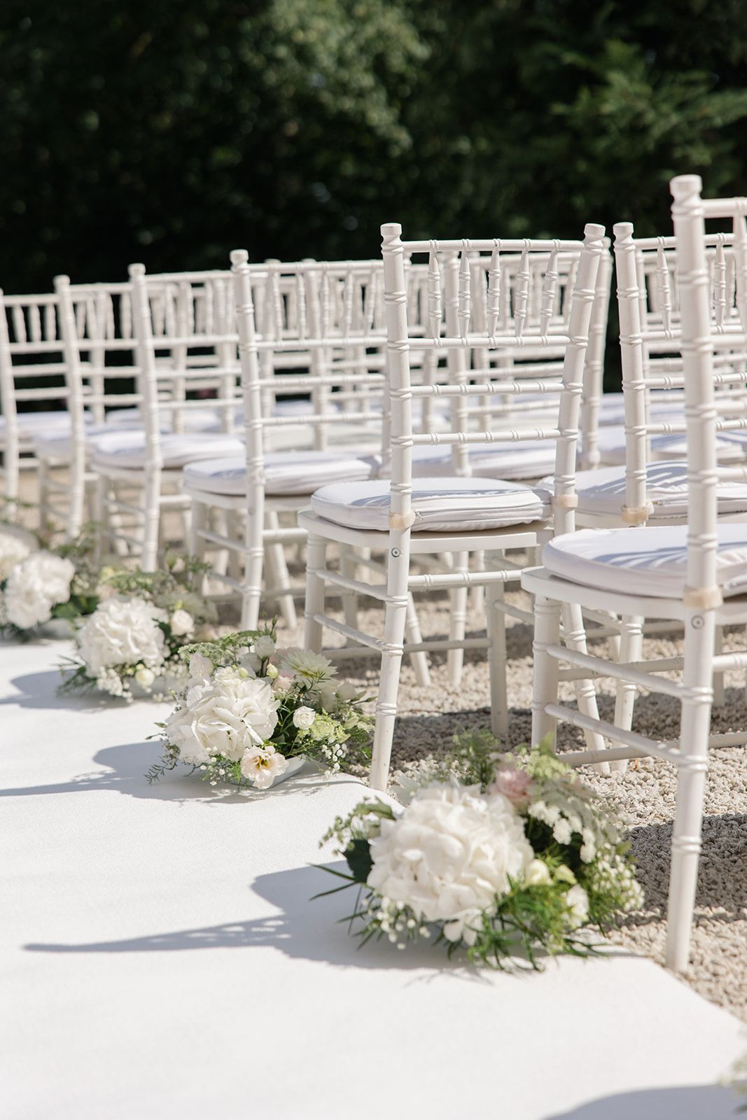 White wooden chairs arranged in rows with delicate floral arrangements adorning the aisle at an outdoor wedding ceremony setup.
