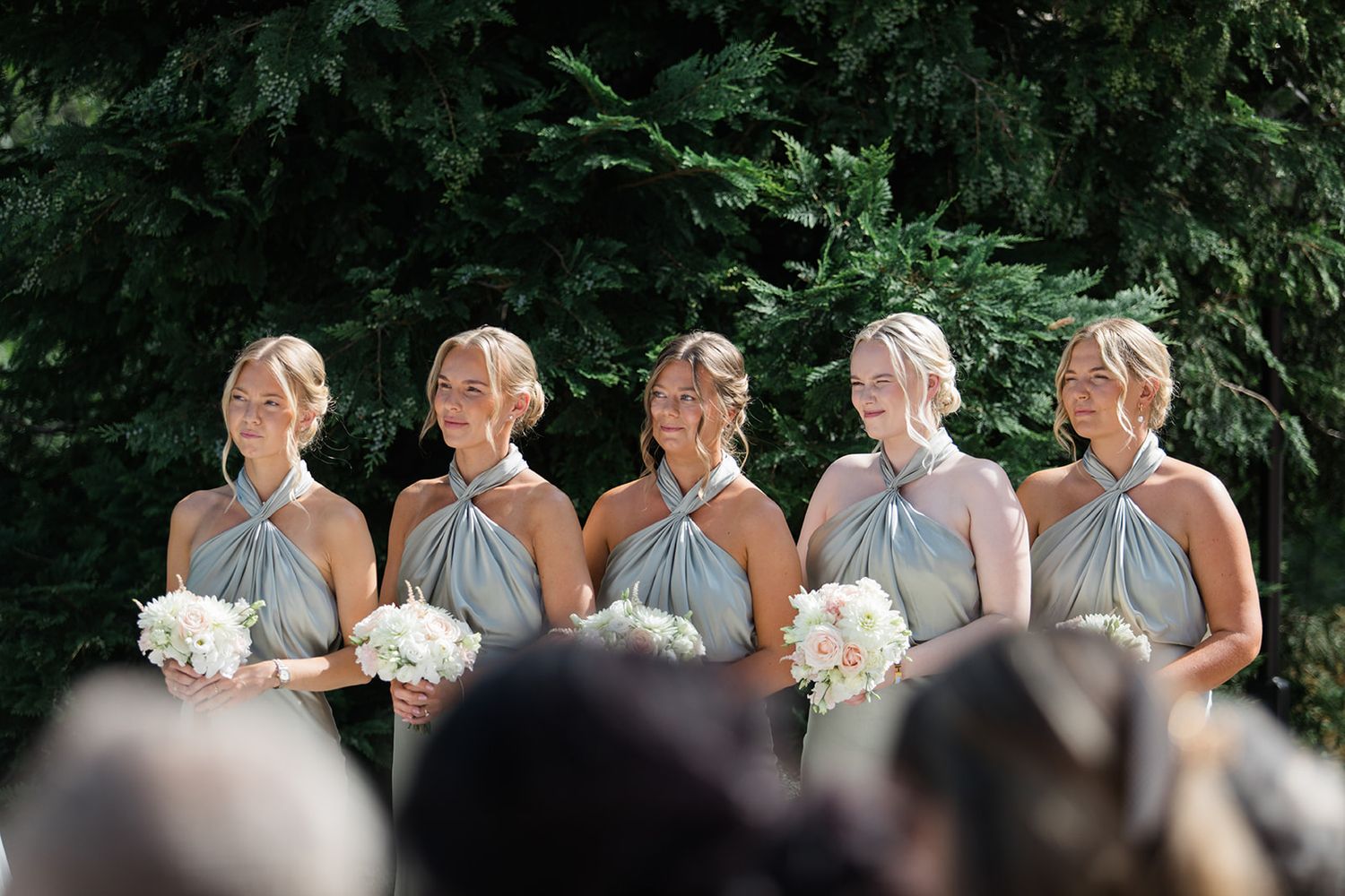 A row of bridesmaids in matching gray dresses stand together holding white floral bouquets in an outdoor setting.