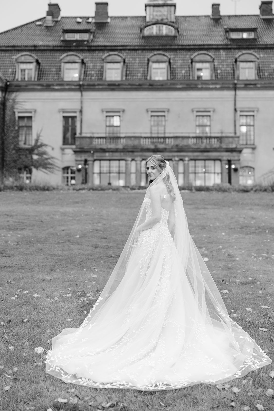 A wedding dress with a flowing lace train and veil photographed in black and white on a grassy lawn.