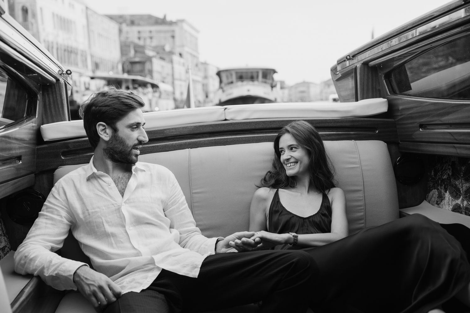 Black and white photo of a couple sitting together in the back seat of a vintage car, smiling.