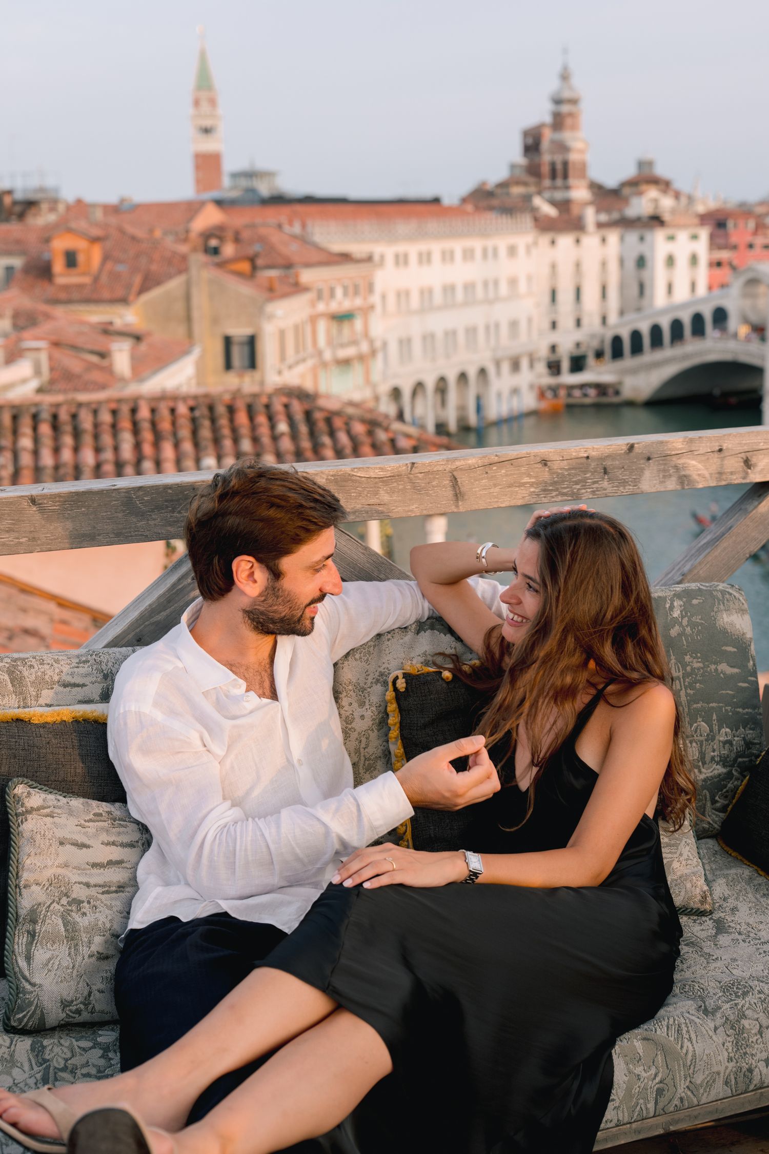 Couple smiling and sharing a romantic moment on a rooftop terrace overlooking Venice canal and Rialto Bridge.