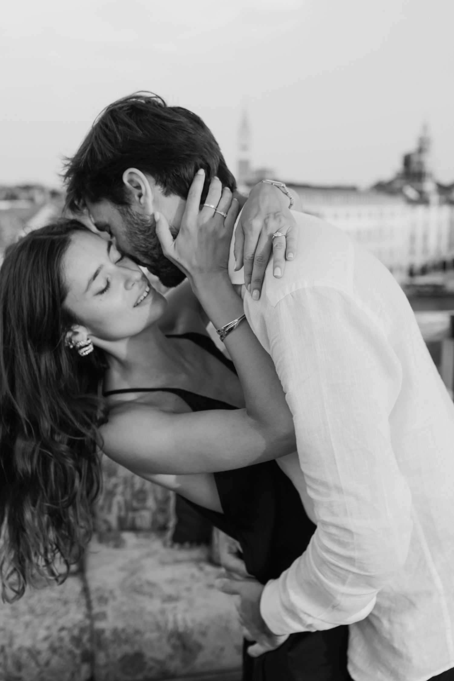 Black and white photo of a romantic couple embracing and kissing outdoors with a blurred cityscape background.