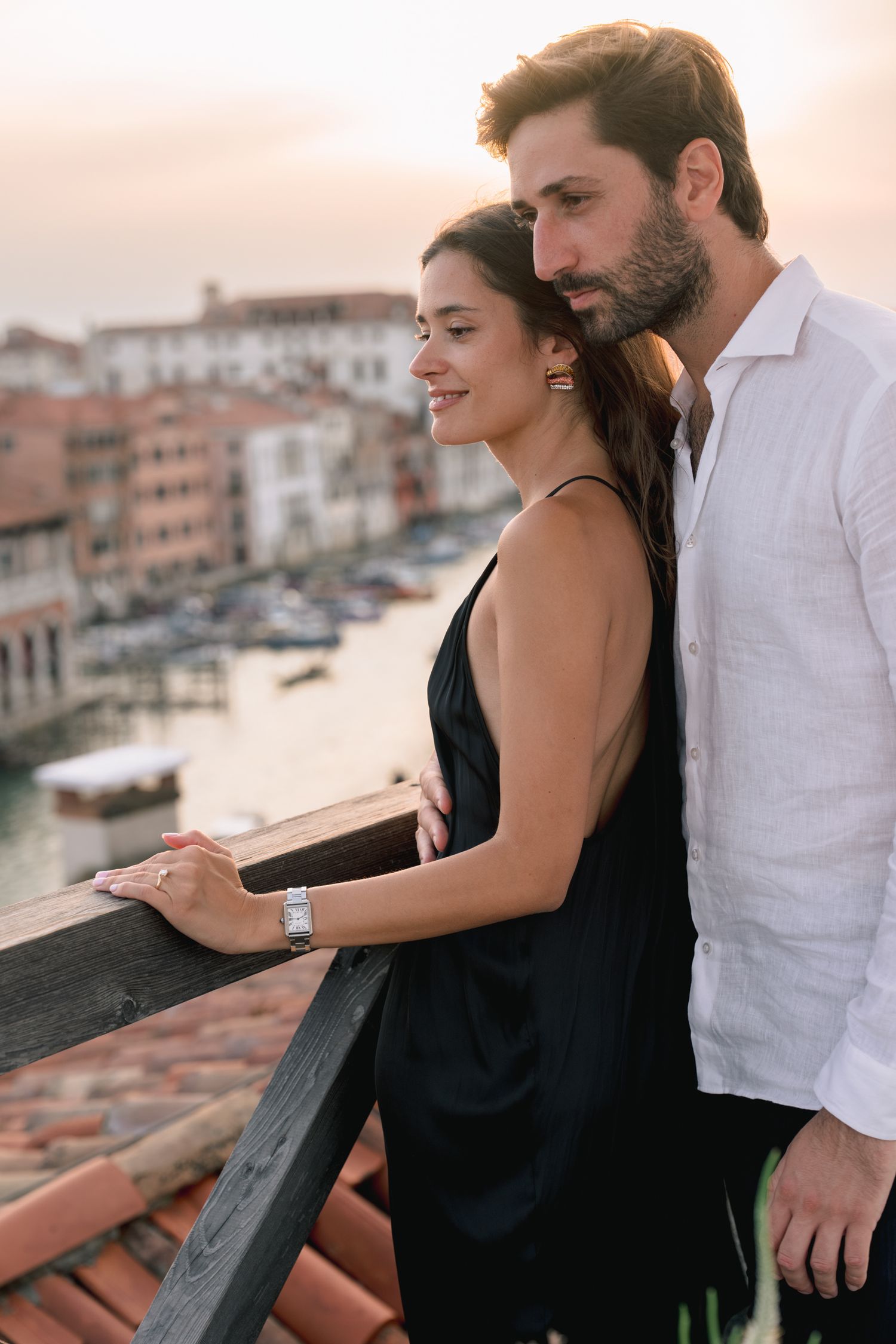 A romantic couple enjoys a stunning Venice canal view from a balcony at sunset, woman in black dress, man in white shirt.
