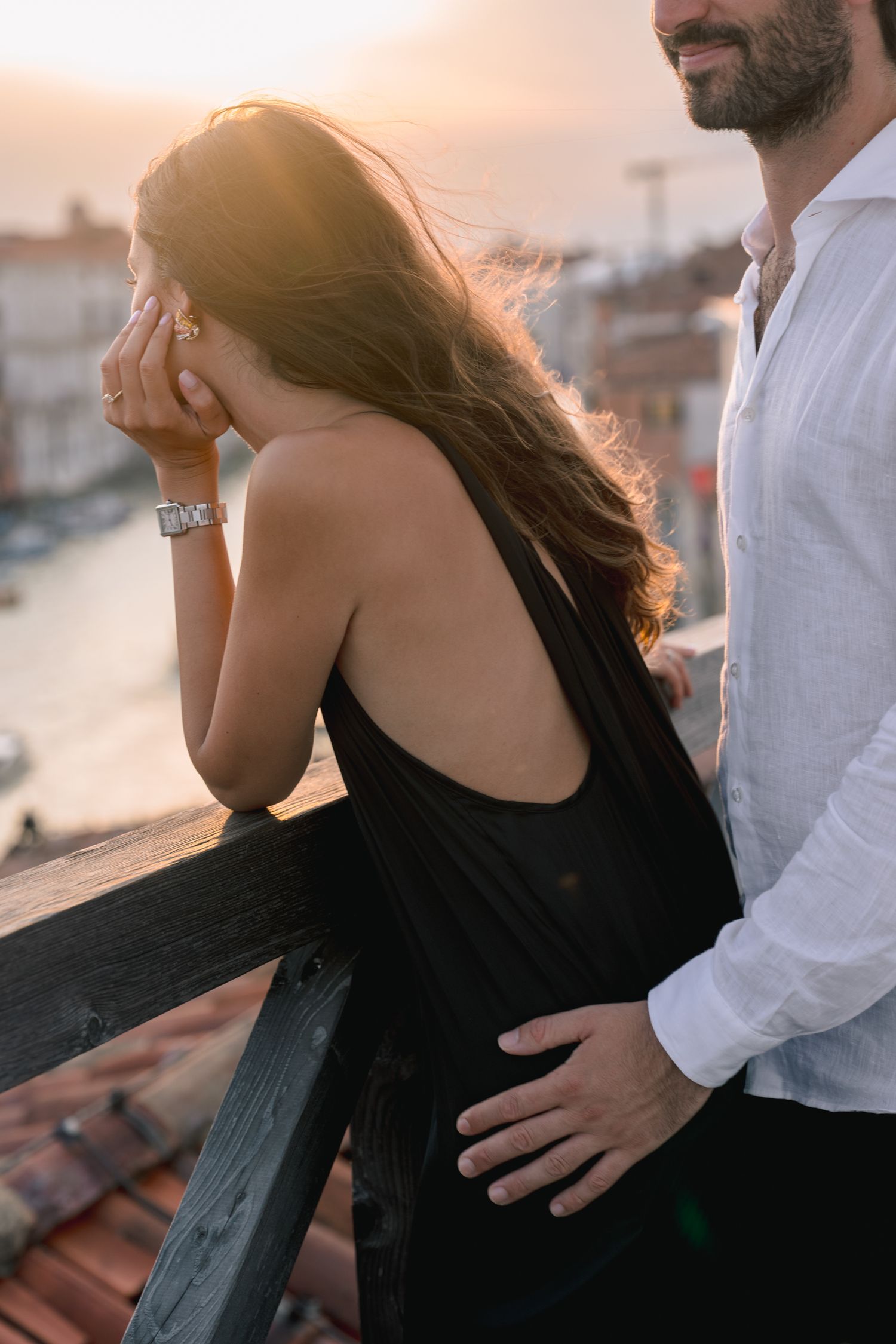 Couple sharing a romantic sunset moment on a rooftop balcony, woman in black dress covering her face.