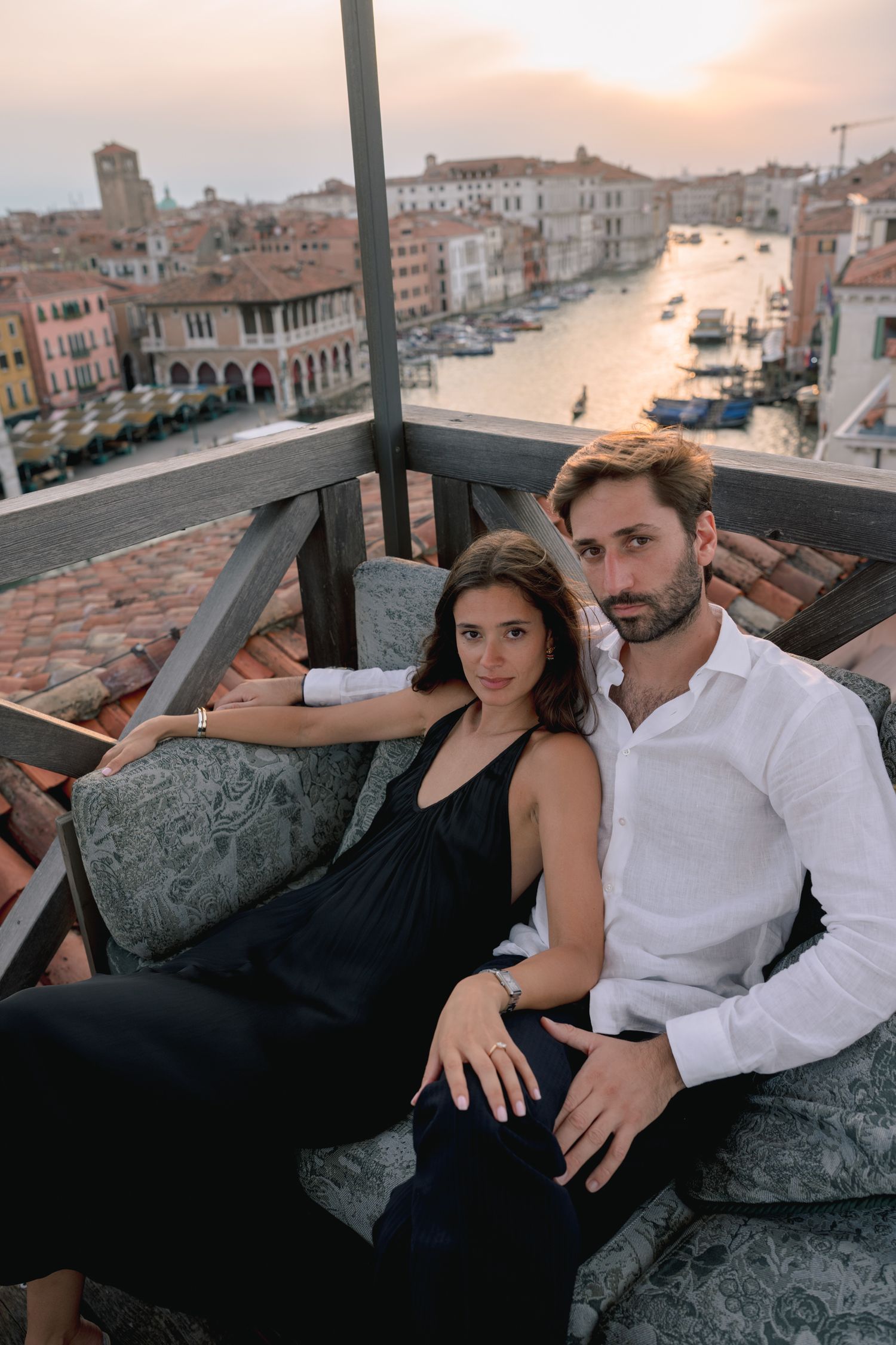 Couple sitting on a balcony overlooking Venice canal at sunset, woman in black dress, man in white shirt.