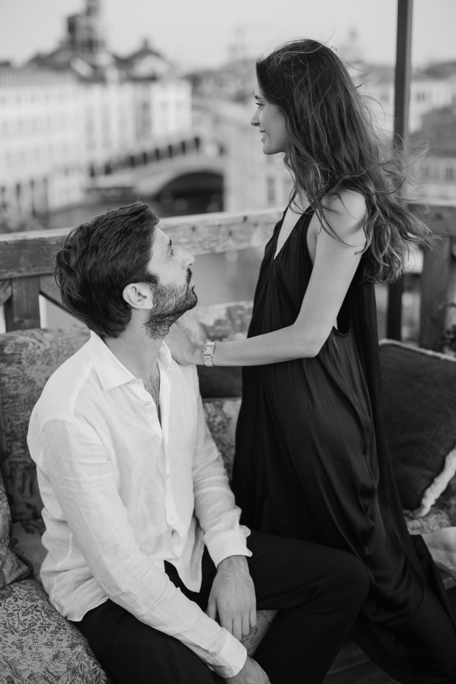Black and white photo of a couple sharing a romantic moment on a balcony with a European cityscape behind them.