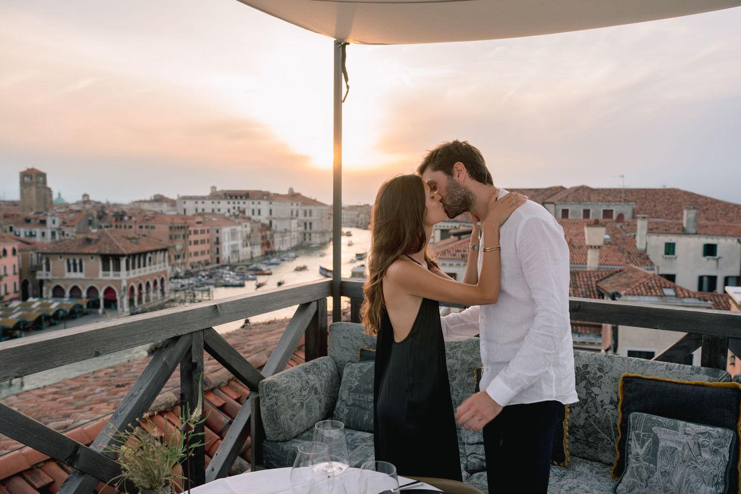 Couple sharing a romantic kiss on a rooftop terrace in Venice at sunset with canal views.