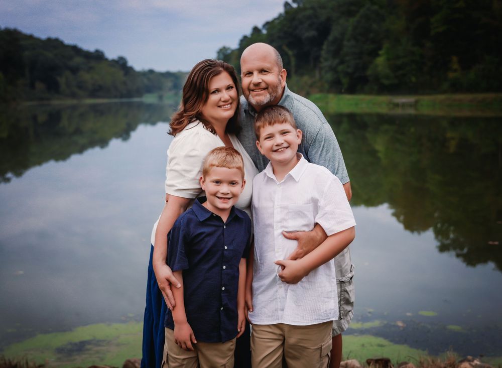 Family of four smiling together by a serene lake surrounded by lush green trees on a cloudy day.