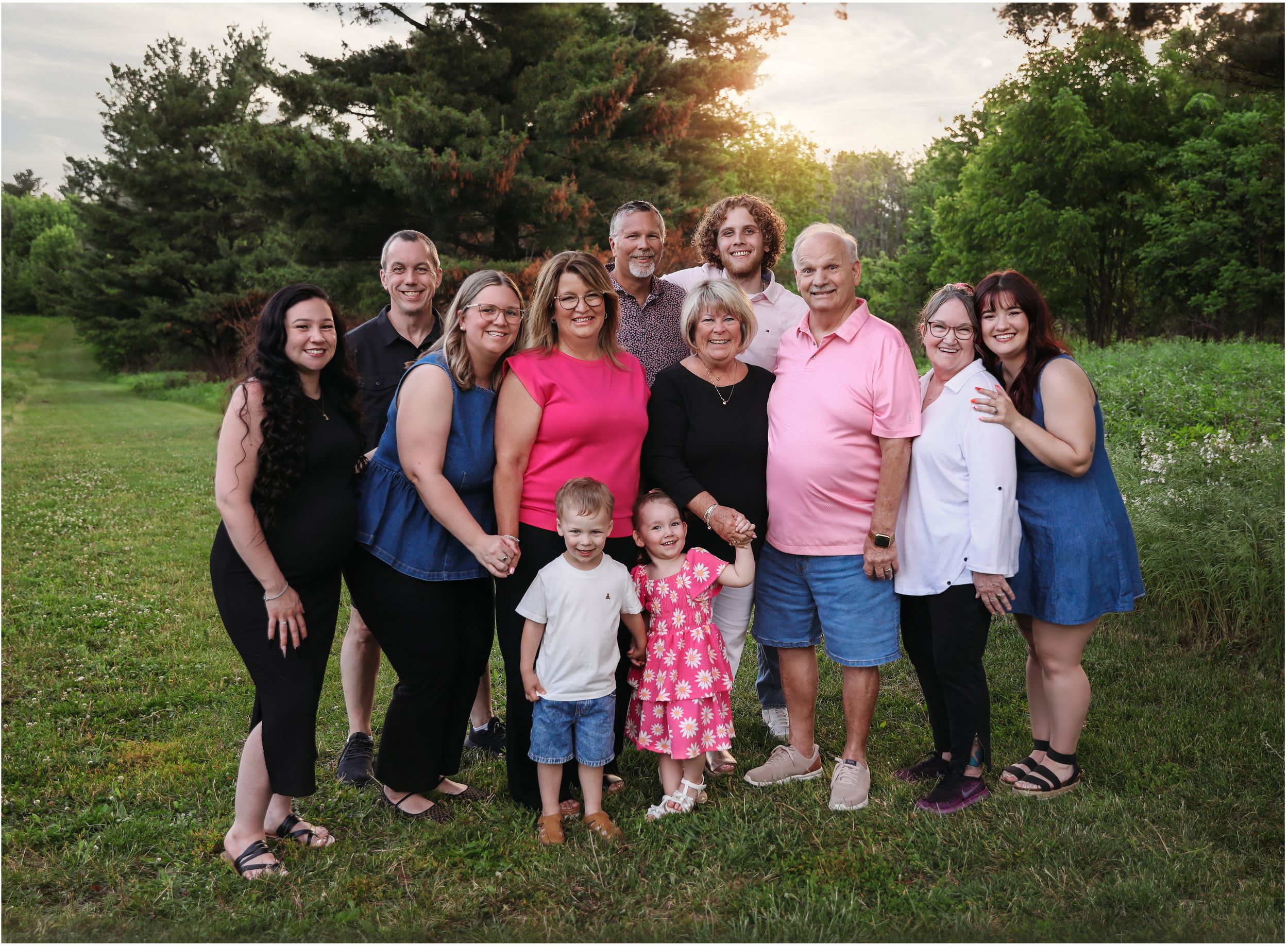 Large family group smiling outdoors on green grass at sunset with trees in background.