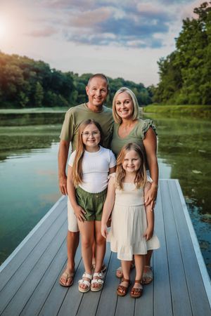 Family standing together on wooden dock by peaceful lake during summer evening.